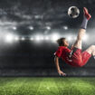 Soccer striker in red uniform hits the ball with an acrobatic kick in the air at the stadium