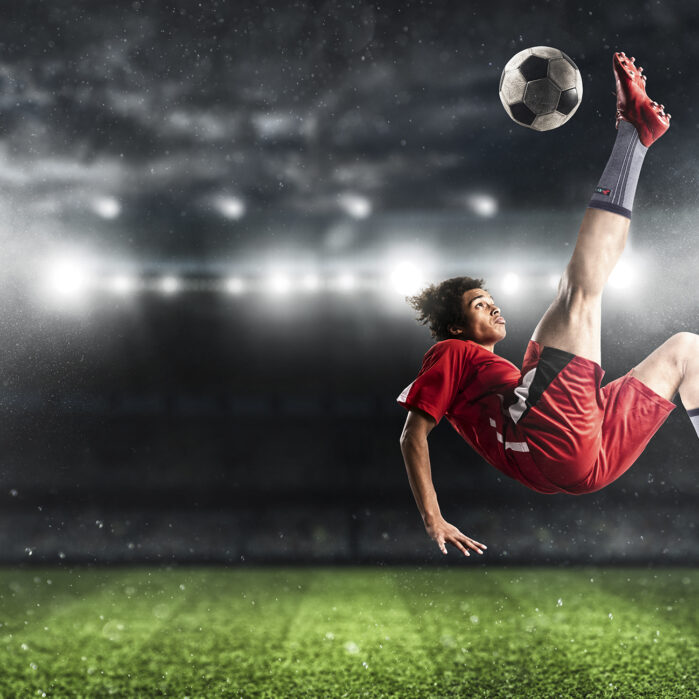 Soccer striker in red uniform hits the ball with an acrobatic kick in the air at the stadium